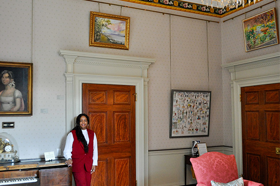 Una mujer vestida de rojo está de pie en una habitación bien decorada bajo un pequeño cuadro de paisaje.