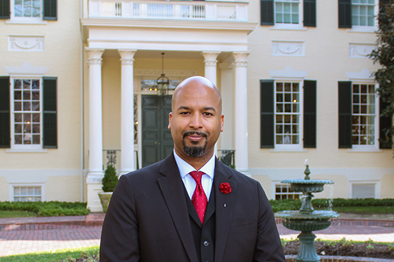 Un hombre bien vestido con un traje de tres piezas con corbata roja se para frente a una gran mansión de estilo federal.