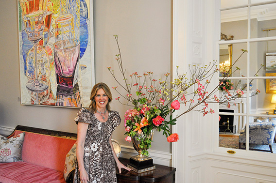 Una mujer está de pie sonriendo junto a un gran arreglo de flores rosas con ramas que sobresalen de él. El arte está en un segundo plano.
