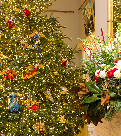 Fondo: Un gran árbol está decorado para Navidad con cálidas luces blancas, adornos de vidrio transparente y ángeles vestidos con túnicas rojas, azules y doradas. Primer plano: En el lado derecho de la imagen, un manto está decorado con rosas rojas y blancas y vegetación con luces blancas cálidas.