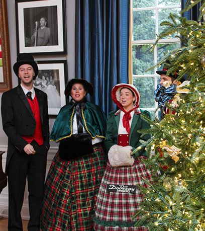 Tres artistas vestidos con ropa de época se encuentran junto a un árbol de Navidad iluminado. Algunos tienen la boca abierta, lo que sugiere que están cantando.