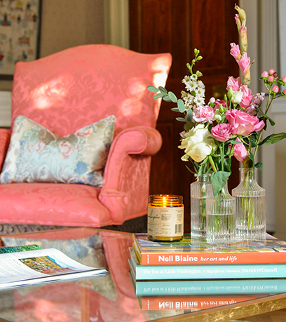 Una mesa de café con tres jarrones de capullos con flores y una vela encima de una pila de libros. Una silla de coral con una almohada decorativa está en el fondo.